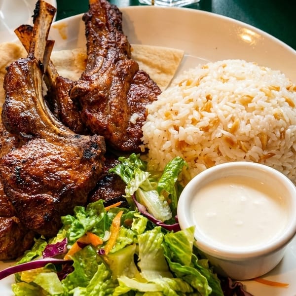 Grilled lamb chops with rice pilaf, fresh salad, flatbread, and a side of white sauce.