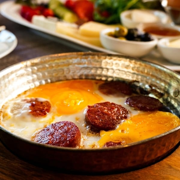A copper pan of fried eggs and sausage on a wooden board, with a breakfast spread in the background.
