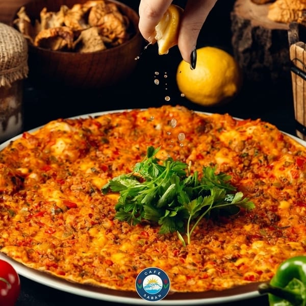 A hand squeezes lemon over a large, savory Turkish lahmacun topped with parsley, surrounded by fresh vegetables.