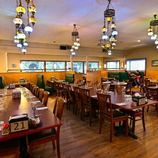 A restaurant dining room with long tables, booths, and colorful hanging lamps, with a fireplace at the back.