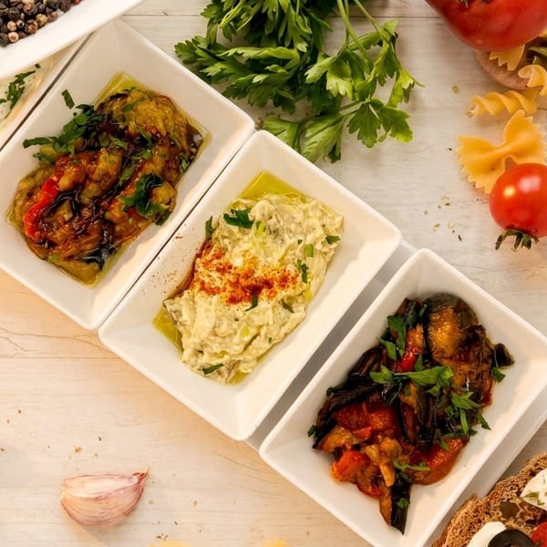 A top-down view of various Mediterranean appetizers in white rectangular dishes on a light wooden table.