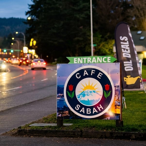 Cafe Sabah sign with an entrance arrow, cars on a wet road, and other business flags at dusk.